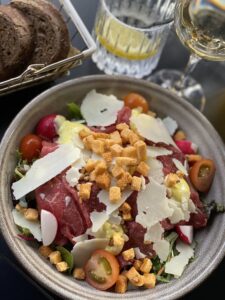 Salat in einer Schale mit Roastbeef, Parmesan und Croutons. Im Hintergrund Brot, ein Glas mit Wasser und ein Glas Wein